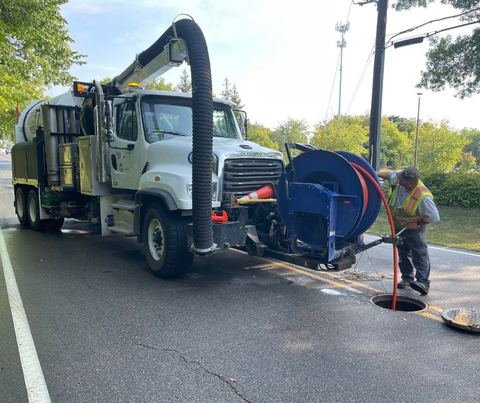 man in bright vest, next to large truck is putting hose into sewer 